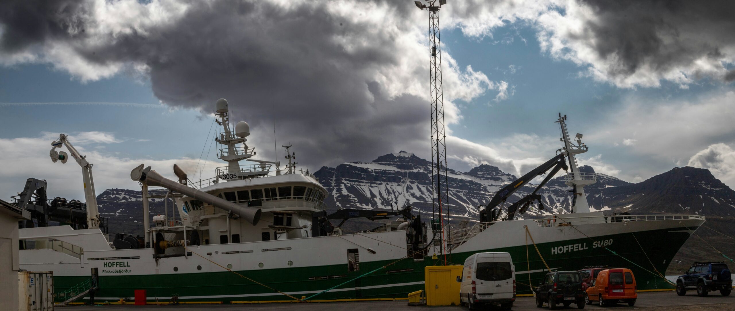 A fishing trawler named "HOFFELL SU80" docked at a harbor in Fáskrúðsfjörður, with the striking Eastfjords mountains of Iceland in the background and an ominous sky overhead.